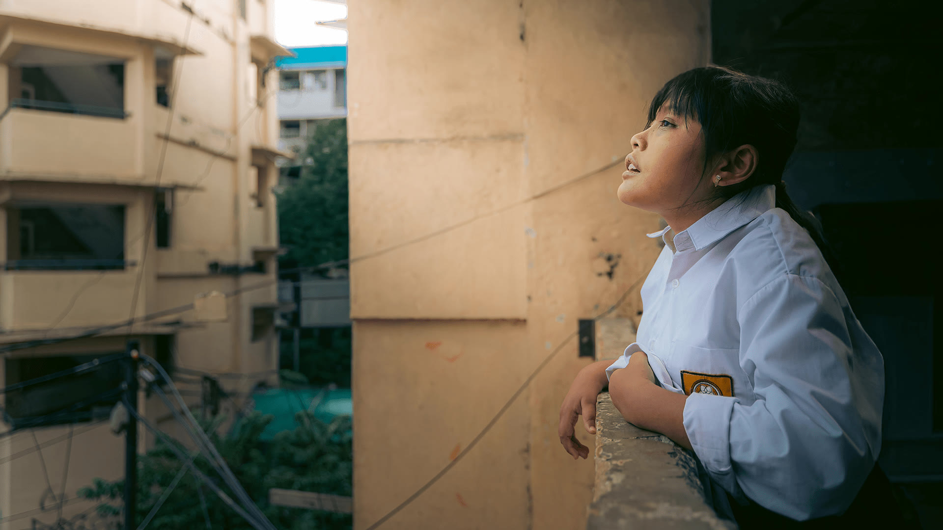 Menina de uniforme escolar apoiada em uma sacada, com prédios do Sudeste Asiático ao fundo